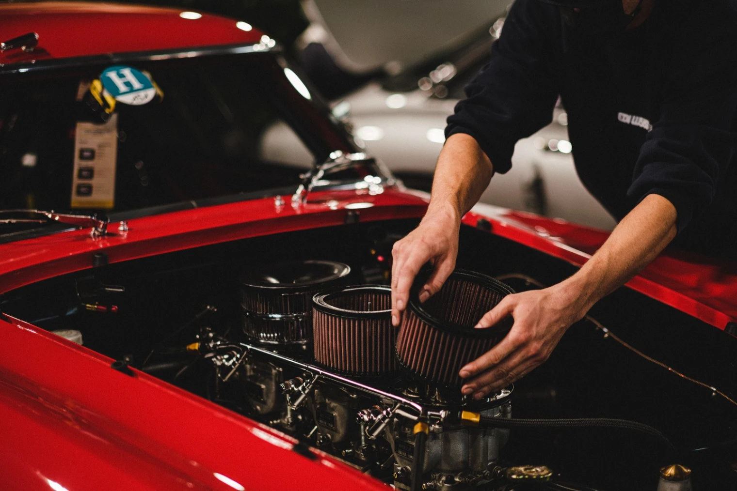 Mechanic placing cylindrical air filters into the engine bay of a red car.